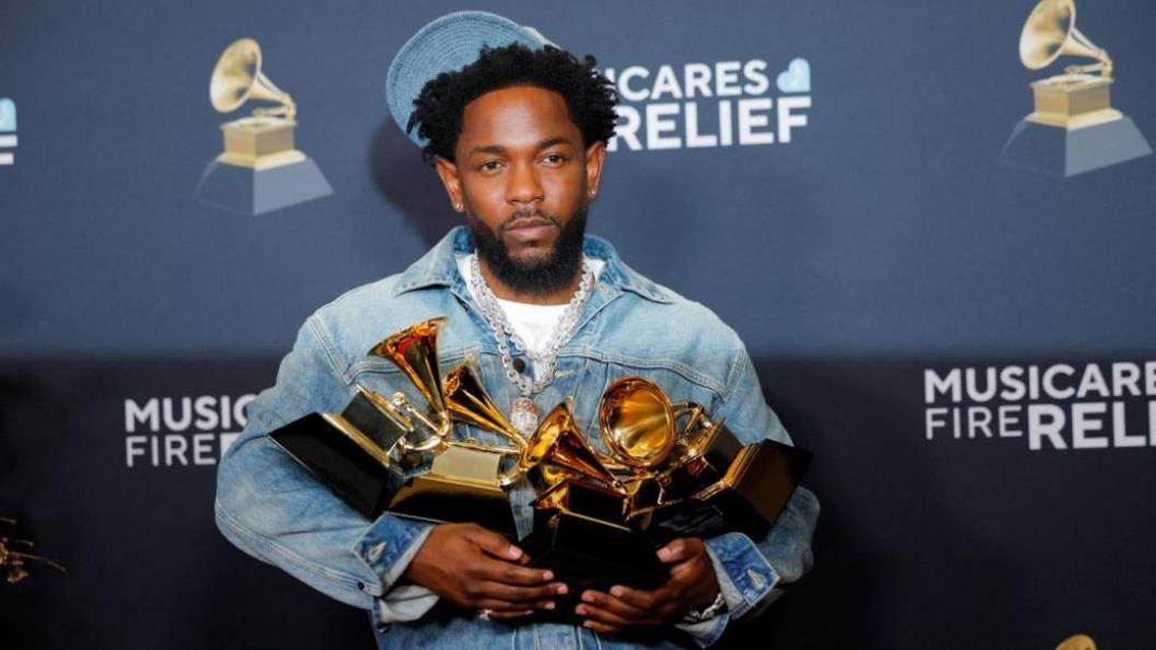 Kendrick Lamar, in blue denim jacket and jeans and heavy silver chain pendant, poses with give golden gramophone shaped awards in his hands in a press room, in front of a black hoarding.