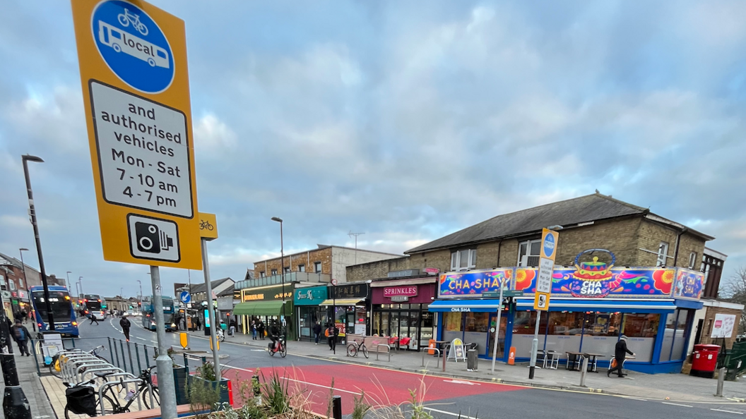 A camera restriction sign on Portswood High Street. There are plants in the foreground with a large yellow sign with a bus and bike symbol and the words "and authorised vehicles Mon - Sat. 7 - 10am. 4-7pm. The road as a read painted stripe across it. You can see shops on either side of the road and a blue bus.