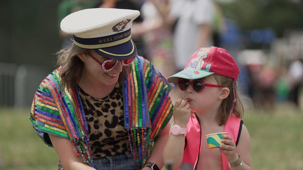 Woman wearing sequined clothing and hat smiles at young daughter whilst she eats ice cream