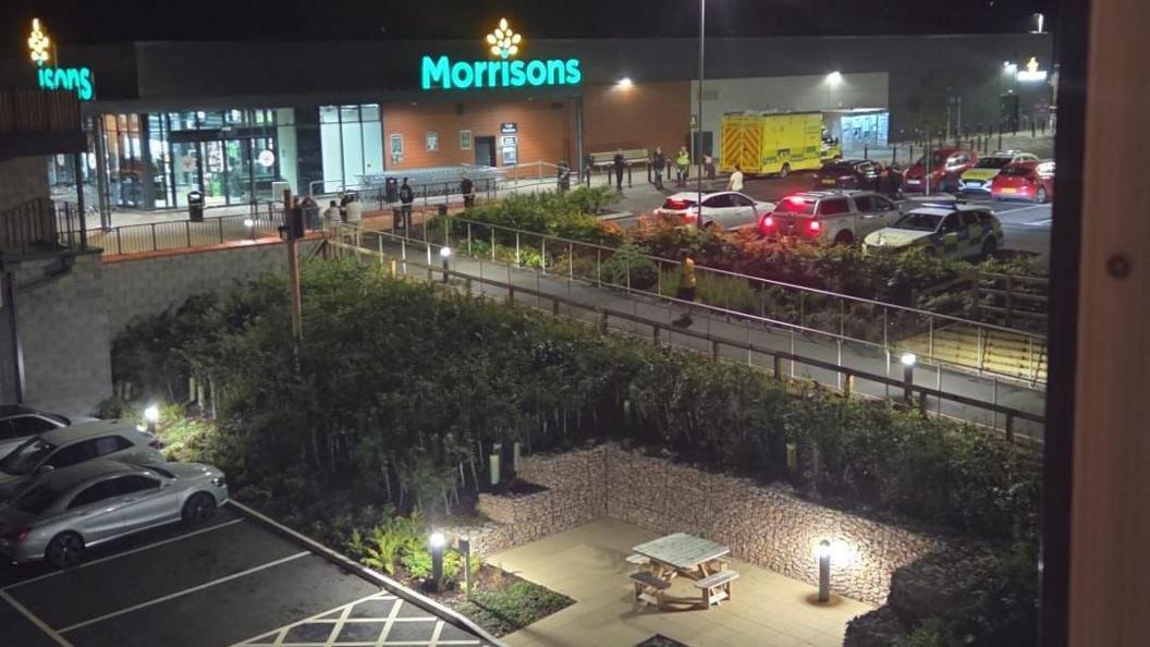 An ambulance and police cars parked outside a Morrisons supermarket along with several other vehicles in the car park. Several people are stood outside the store. It is night time and the supermarket's logo is lit up.
