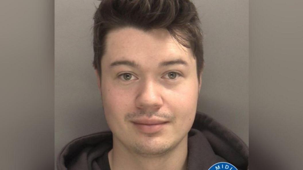 The face of a young man with short dark hair and a dark hoodie against a grey background