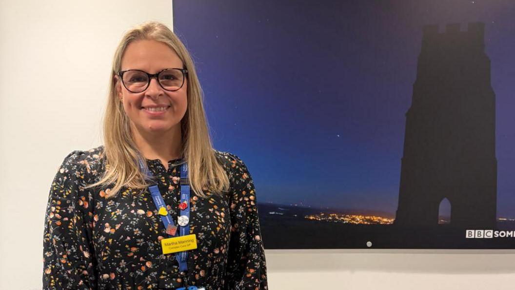 Dr Manning is standing on the left of the frame, wearing a blue NHS lanyard and yellow name badge over her dark, autumnal coloured dress. She's got shoulder length blonde hair. She is standing next to a picture of Glastonbury Tor.