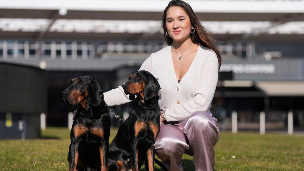 A woman with a white buttoned top and mauve jeans squats on a grass field next to two dogs. The dogs have a mix of black and brown fur and look to the left. The woman has long dark brown hair, past her shoulders.