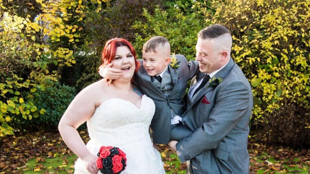 Leigha Cartmell with red hair and white wedding dress, holding red flowers. James Cartmell in a grey suit holding Jay who is smiling.