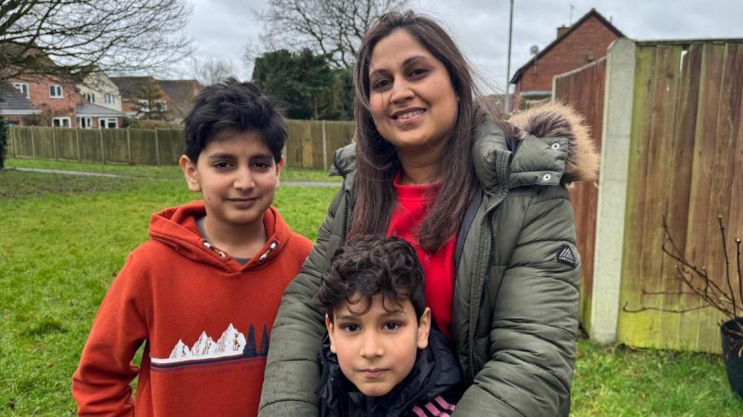 A family picture of a mother in a thick green coat and her two young sons in jumpers standing in a local park surrounded by wooden fence panels and some trees. 