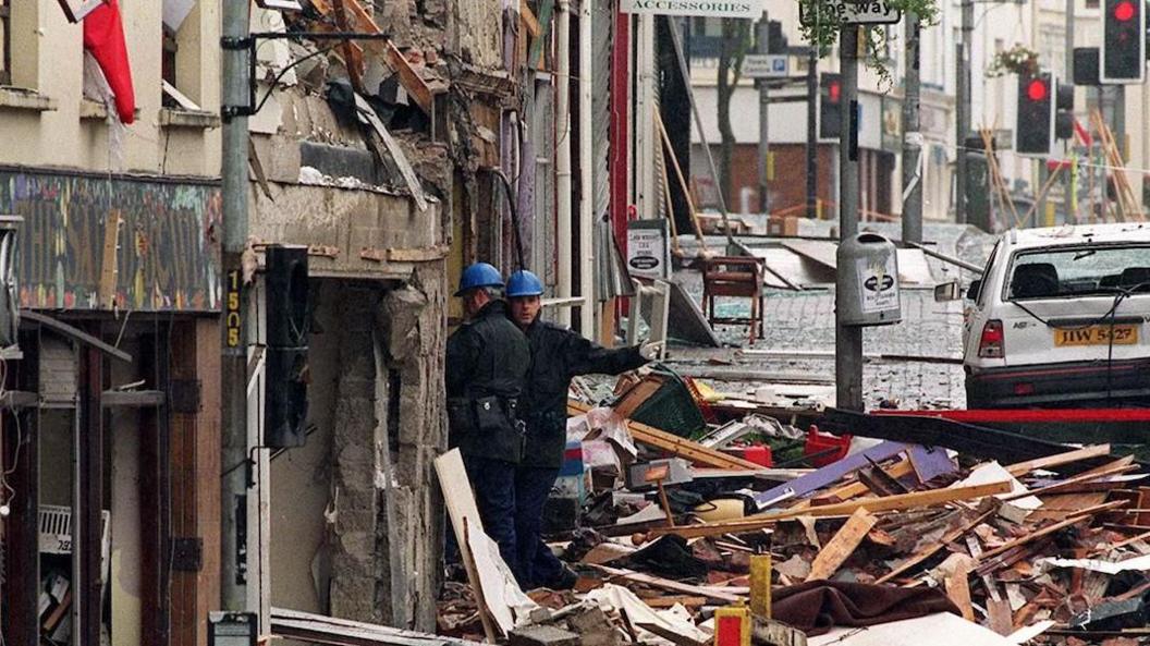 Two men in blue uniforms and blue hard hats examine the rubble in the street. The buildings and shop fronts are destroyed and a white car has its windows blown out.
