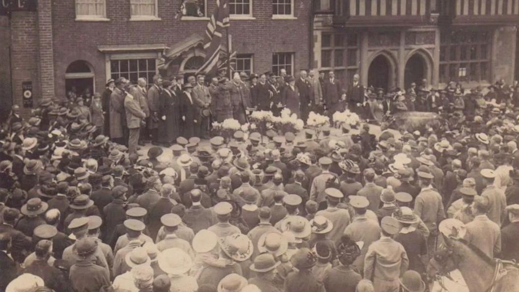 A black and white image of a two-minute silence in Farnham on 1 May 1916 with crowds of people and dignitaries with flags on stage in front of a big building
