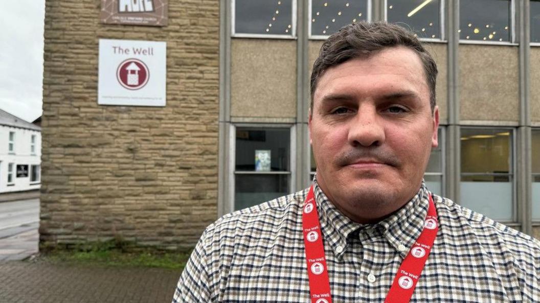 Danny Woodburn standing outside an office block which houses The Well. He is a thick-set man with short, neat dark hair with a side parting and slight wave. He has the shadow of a moustache and wears a red lanyard over his check shirt. The ground and first floor of the offices can be seen behind him. It has stonework on the left, on which signs for Hope Hub and The Well are attached. The main bulk of the building consists of rectangular windows with little horizontal gap between each. The windows on each floor have a larger, beige concrete panel separating them.