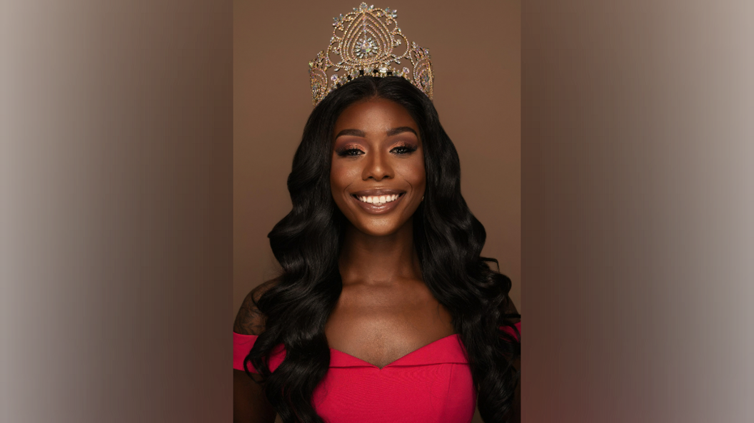 A black woman smiles at the camera, wearing a gold crown and pink dress. She has long black hair past her shoulders.