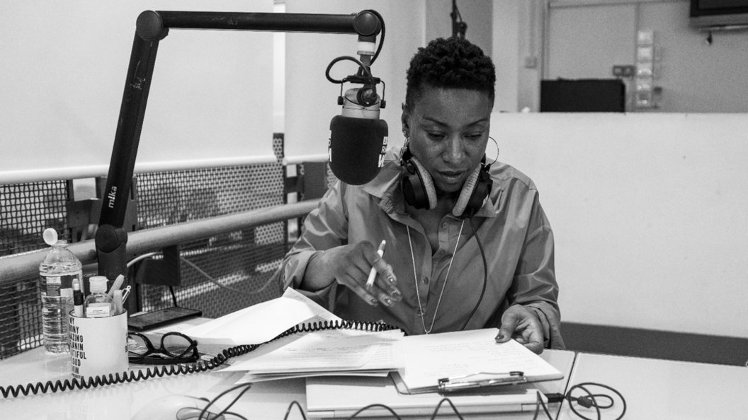 Nikki is at a desk with paper and notes in front of her and a microphone. She has headphone on and is looking at her notes in this black and white photo.