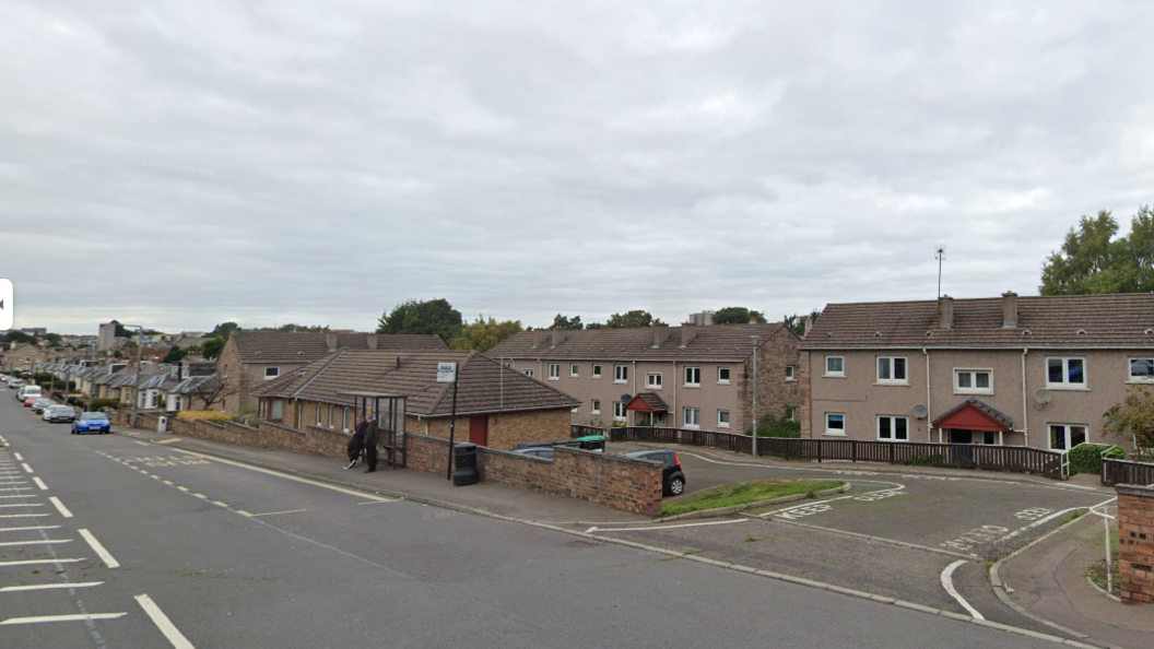 A road lined with brown houses. There is a bus stop and some parked cars