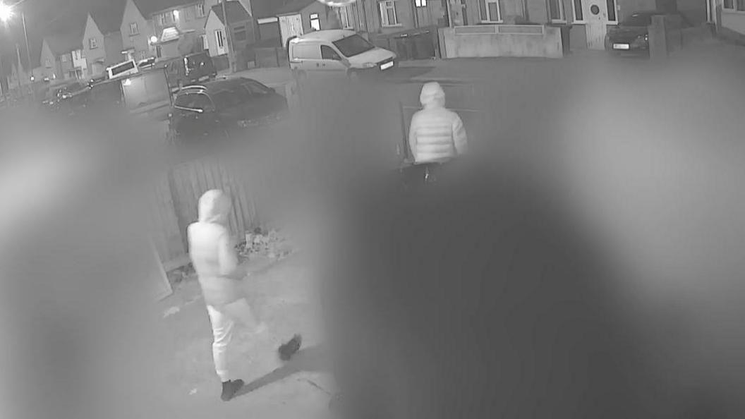 Black and white CCTV footage capturing Max and Mason wearing tracksuit bottoms and winter coats with their hoods up. They are leaving the front yard of Mason's home and walking through the gate onto the pavement of Ilminster Avenue.