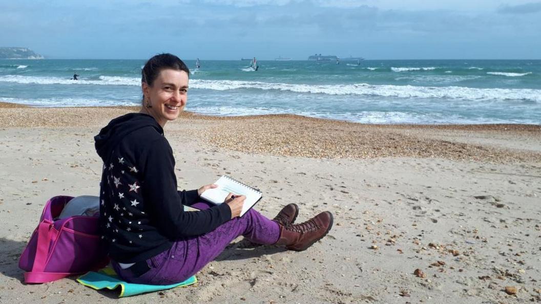 Nicki Greenham sitting on a beach on a cloudy day, turning around to smile for the camera. She is sitting on a blue towel and is holding a notepad and a pencil. There are a few boats in the sea behind