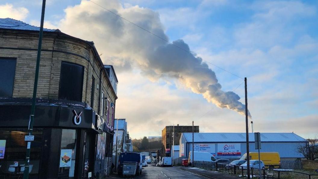 A street scene with various shops on one side and industrial buildings on the other. Dominating the skyline is a tall chimney with a huge plume of white smoke coming out of it and stretching across the sky.