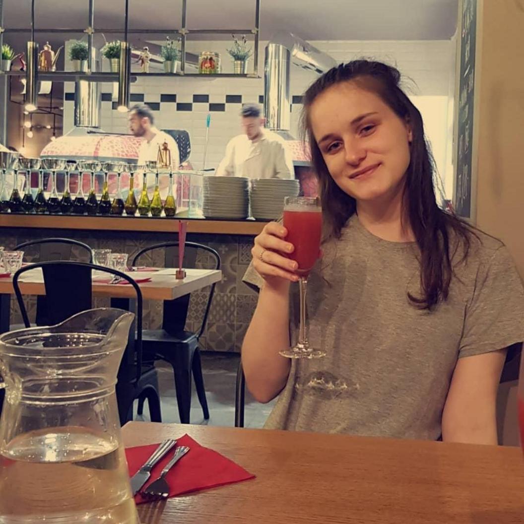 Cerys Lupton-Jones sitting in a restaurant. She is smiling at the camera and holding up a tall wine glass with a red drink in it. She is wearing a grey short and has long brown hair over her shoulders. Behind her two chefs can be seen working in an open kitchen