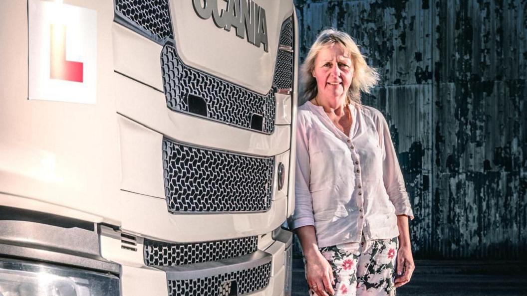 Sue Howe, a white woman, stands at the front of a large truck with a Learner Driver L plate on it. She is wearing a white shirt and trousers with a flower pattern on them