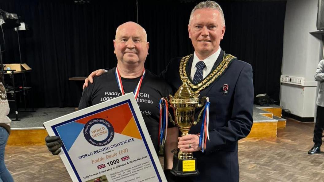 A bald man is in a black T-shirt, with black gloves and a blue, white and red medal strap around his neck. He is holding a large certificate titled "world record certificate". it reads "Paddy Doyle, 1000, 1000 fitness endurance challenges and world records". He is standing next to the Sutton Coldfield mayor, who has a suit on as well as his mayoral chains. He is holding a trophy.