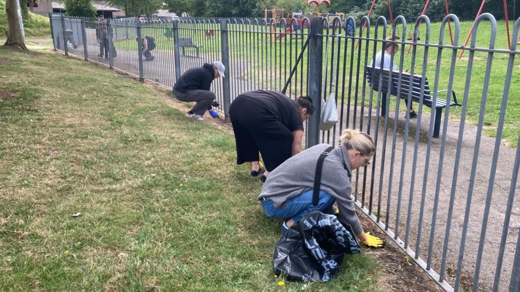 Two women bending down and clearing up rubbish from a grass area next to a fence. One woman has blue jeans and a grey jumper and yellow gloves, while another has black trousers and a black top. A man is also helping and is bending down in the background wearing a grey cap.