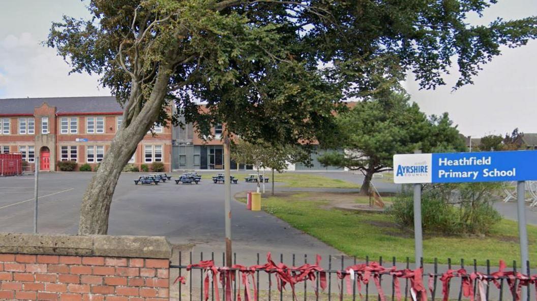 Healthfield Primary School is a red brick, two storey building with a large playground in front of it. The playground has several picnic tables with some patches of grass with a couple of trees. A blue sign reads "Heathfield Primary School". 