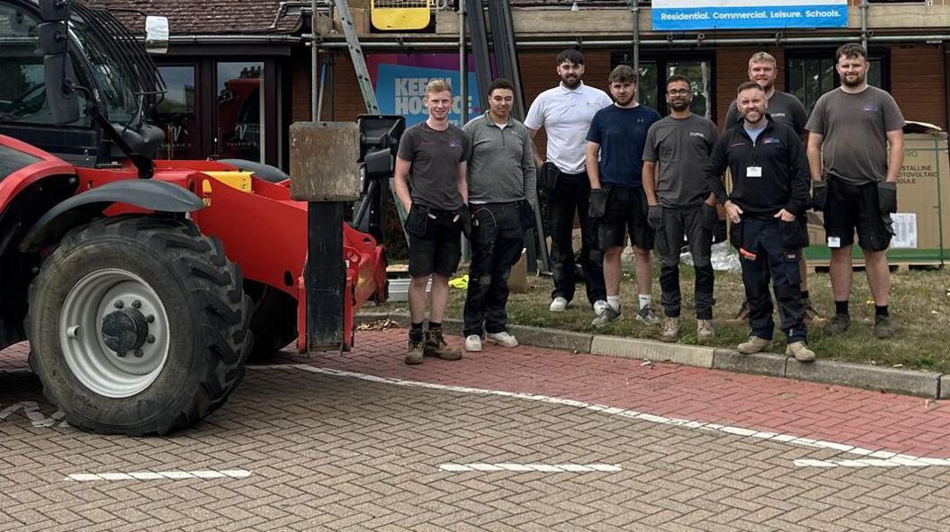 The front of a large red fork-lift truck parked in front of Keech Hospice. A group of eight men stand in a group looking at the camera, wearing workman's trousers or shorts and T-shirts. Behind them you can see the scaffolding against the building.