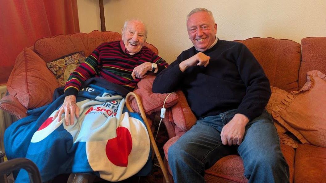 Two smiling men sat down next to each other. On the left is an older man, with balding light grey hair. He is wearing a striped coloured jumper and has a blanket over his knees with a walking stick resting to the side. On the right is a younger man, wearing dark blue jeans and a dark navy jumper. 