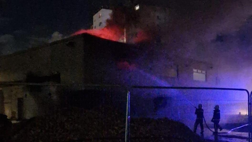 Firefighters can be seen spraying water at a two-storey white building - the Low Grange social club. It is night-time, but the picture is illuminated by orange flames coming from the roof and the blue lights from the emergency vehicles.