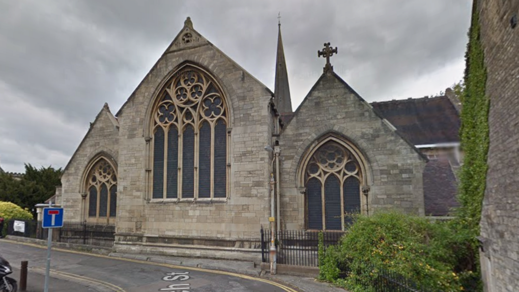 The frontage of St Laurence Church in Stroud as seen on a cloudy day. The church has three separate frontages with large windows visible with the tower in the background. A road, yellow lines, a no-through road sign and a green creeper growing up a wall can also be seen on a wall.