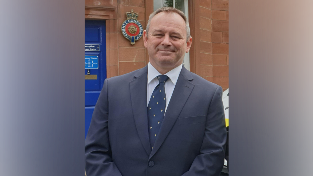 David Allen with short brown/grey hair, wearing a navy suit, white shirt and navy tie, stands outside of a brown brick building.