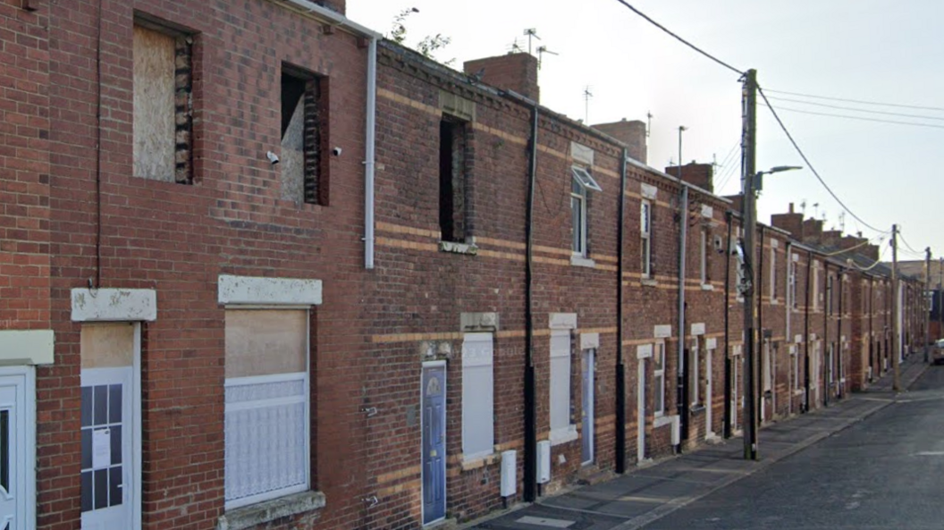 A street of vandalised terraced homes with boards up in the windows and pictures of fake front doors stapled on to the fronts of boards over the front doors.