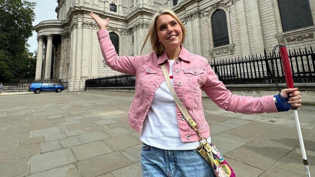 Claire Sisk standing outside St Paul's Cathedral in London, she is standing on a pavement, with both her arms outstretched. She is holding a walking cane, with a red top. She is wearing jeans and a white T-shirt, a pink denim jacket and has a bag across her body. A blue car and St Paul's Cathedral can see been behind her. 