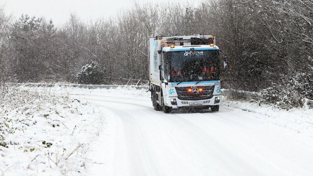 A white and blue bin lorry with flashing lights drives on what appears to be a lane of a roundabout that is covered with snow and while snow falls 