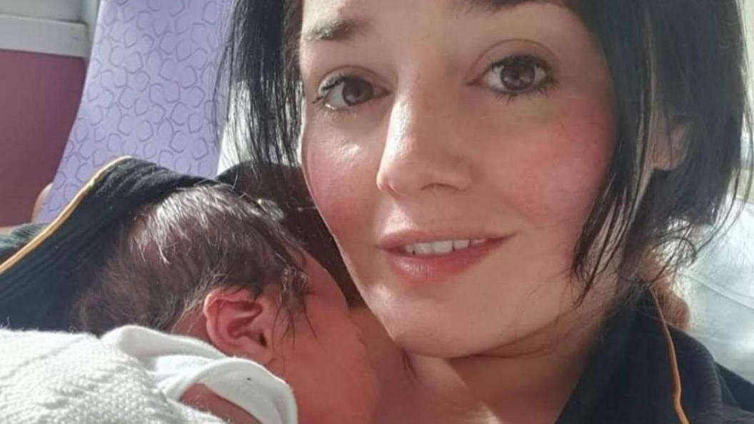 Niki Cotson cradling her newborn son while lying on a hospital bed. Both have dark hair, with Niki's being in a short bob cut.