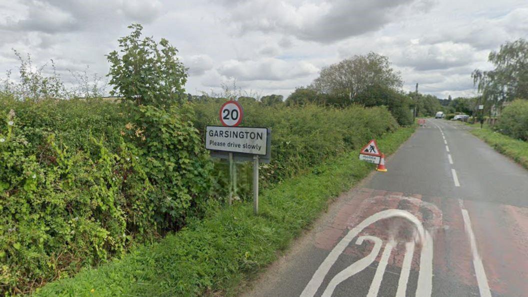 A road view of Oxford Road into Garsington. There is a sign with a 20mph speed limit. There is a roadworks sign and a cone on the road.