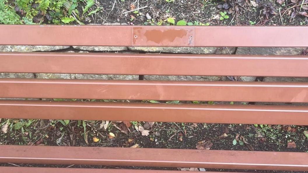 A close-up of a brown wooden park bench with two small holes drilled into it and a rectangular outline and some damage that looks like it has been made from something being pulled off. 