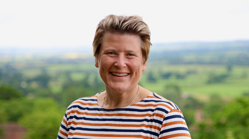 Sarah Dyke wearing a blue and orange striped t-shirt and a silver necklace. She has short blonde hair and is smiling at the camera. In the background there is a blurred landscape view looking over green fields and trees. 