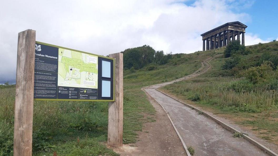 A swerving path leading up to Penshaw Monument, which resembles a Greek temple with stone pillars. There is a National Trust sign at the bottom of the hill with a map and information about the site.