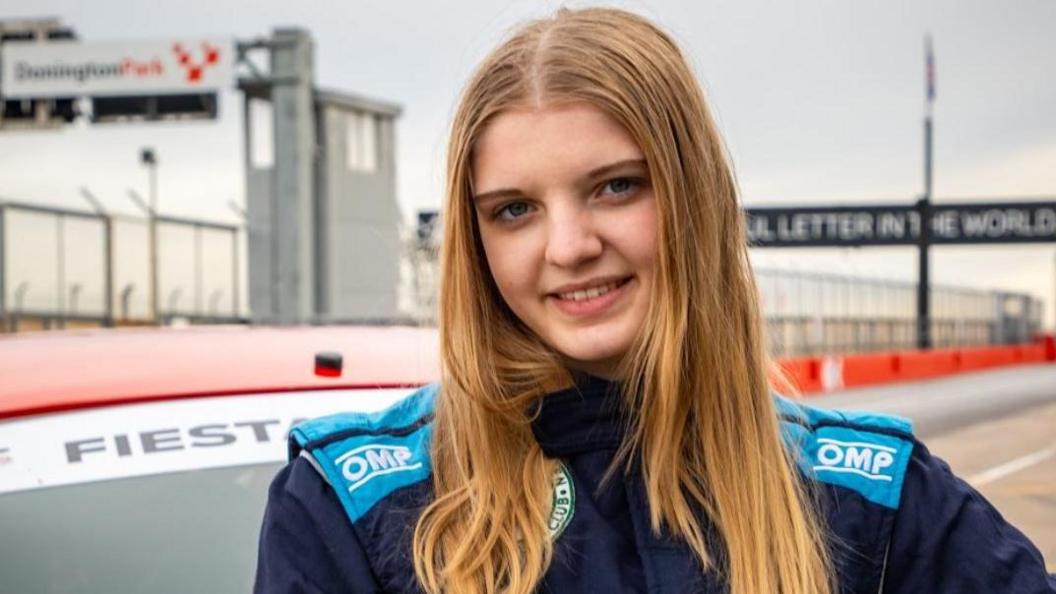 Daniella Sutton has long, straight hair and is wearing a racing suit. She is stood next to a car on a race track and is smiling at the camera.
