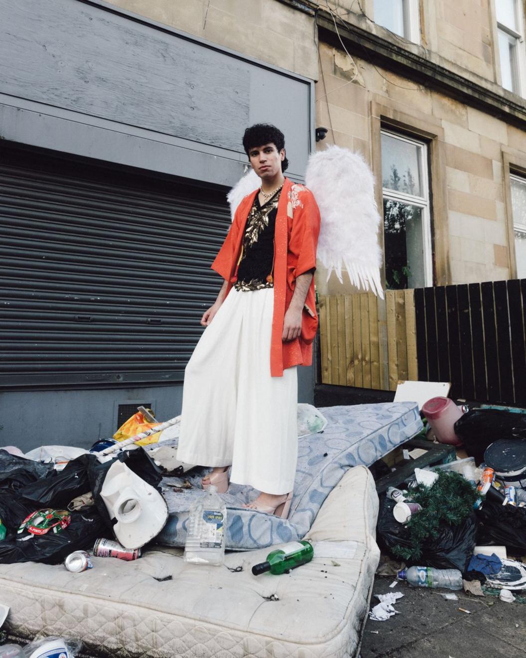 Jacob Alon, wearing a pair of white feathered wings, stands in a back yard that's been used for fly-tipping mattresses, old clothes and other household waste