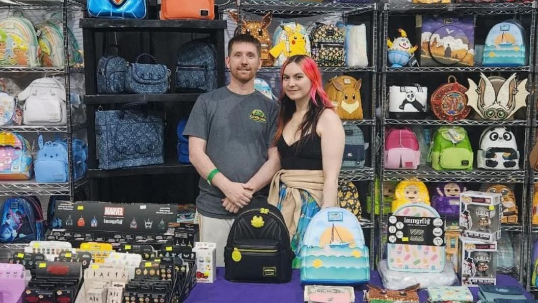 Two people - one a man and the another a woman- stand behind a large table. On the table and on shelves behind them are rows of loungefly bags, of various colours and designs.