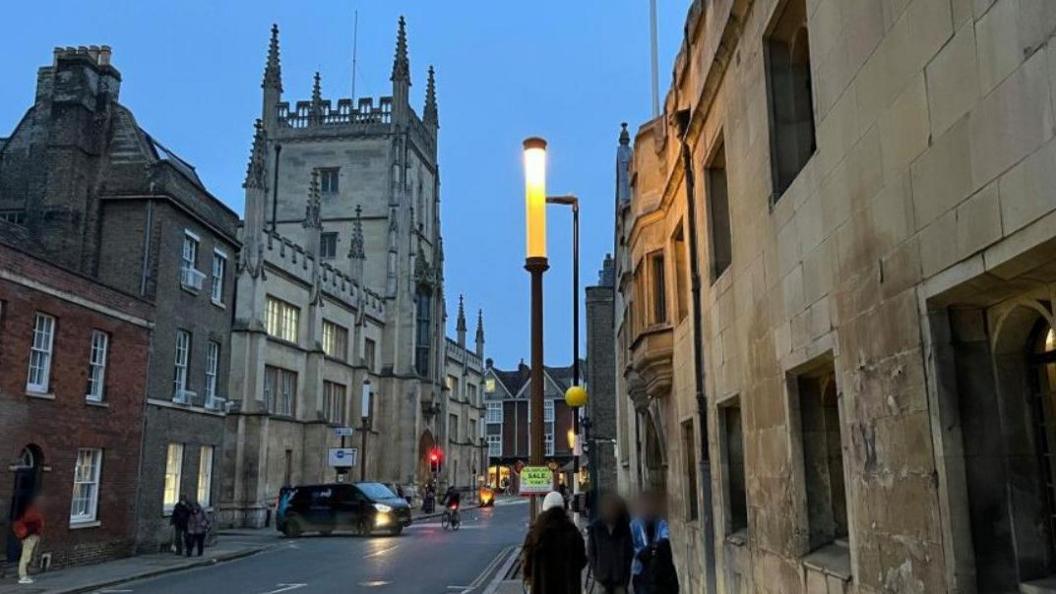 A lamp on a street in Cambridge at dusk. It is tall and thin and lit up. There are historical buildings on the street and pedestrians walking on the pavements. There is a van and a cyclist in the road