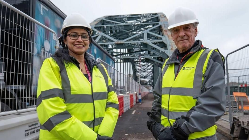 Newcastle City Council councillor Juna Sathian and Gateshead Council councillor John McElroy standing on the Tyne Bridge during the repair work. Both are wearing council branded hi-vis jackets and white hard hats. Wire fencing is on either side of them. The Tyne Bridge is behind them and rust can be seen among its green paint.