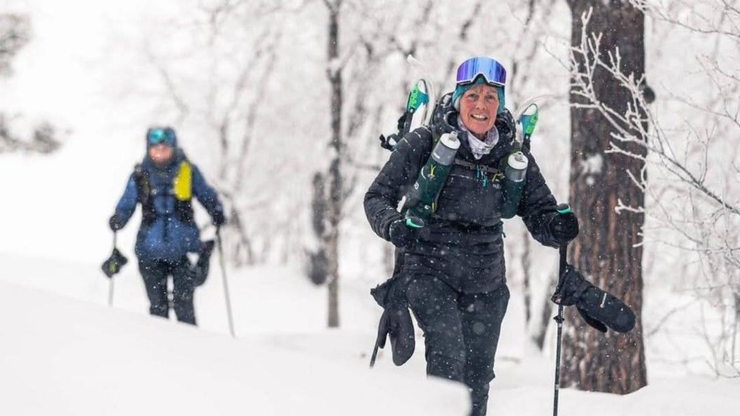 Two warm clothed women with poles, ice goggles, gloves and rucksacks, trudging through a snow covered forest landscape