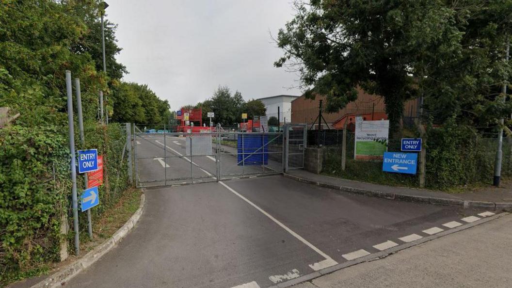 The entrance to Yeovil Recycling Centre. A road leads to metal gates with the recycling centre beyond them.