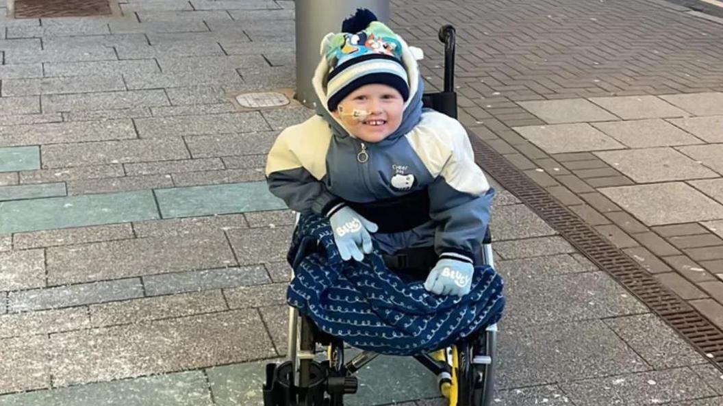 A boy with a blue and white coat, blue gloves an a hat with Bluey on it sat in a wheelchair on a pavement with a blue blanket on his lower half.