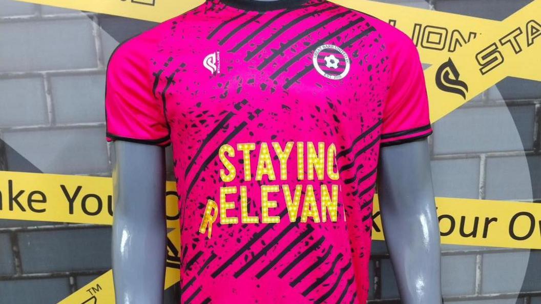 A pink and black football shirt with a yellow logo in the centre of the shirt that reads "Staying Relevant"