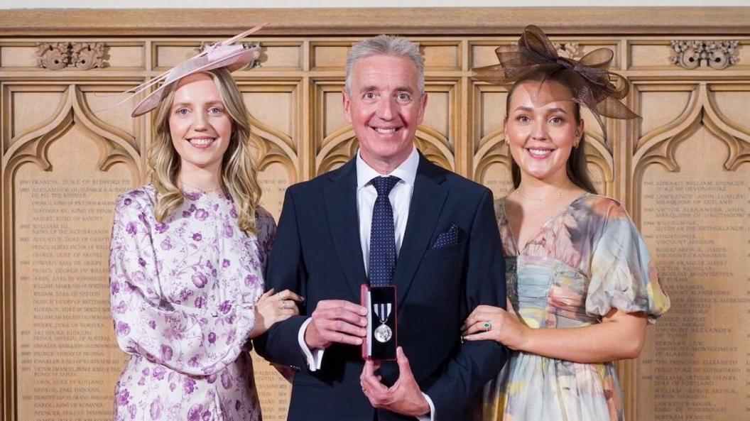 Jim has short grey hair and is dressed smartly, whilst holding a medal. Each of his daughters cling on to his arms.