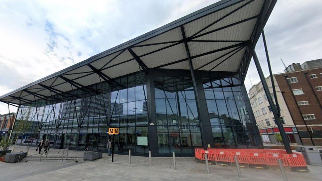 Sunderland train station. It is a tall glass building. There is a Tyne and Wear Metro sign in front of the entrance. Several people are walking past.