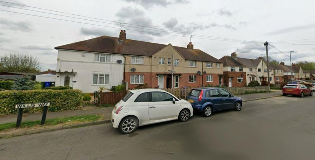 A row of blocks of two-storey houses. The nearest block has a brick ground floor and light-brown painted upper floor. There is a white car and blue car parked on the road outside the houses. There is a green hedge to the left of the picture. A white roadsign indicates WILLIS WAY in black lettering.