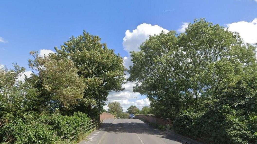 A road, with trees overhanging from either side, and a pavement on the left hand side.