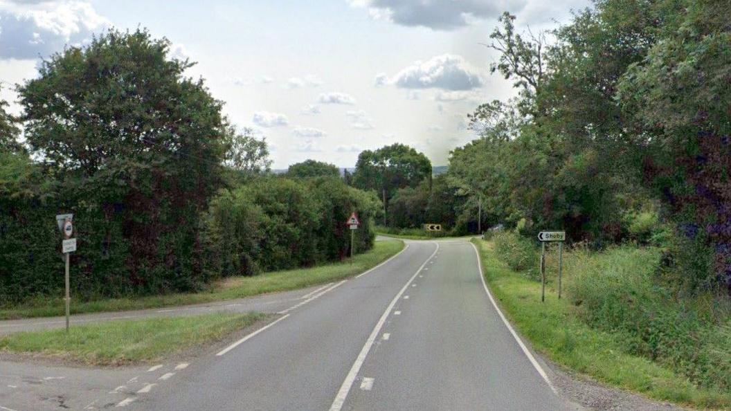 A general street view of the A6006 in Leicestershire near Shoby. There is a small signpost on the right of the road, opposite a junction on the left, which reads 'Shoby'